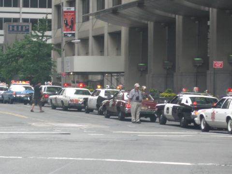 1991 Ford LTD Crown Victoria - long row of vintage police cars is parked along a city curb beside an office building as pedestrians walk past.