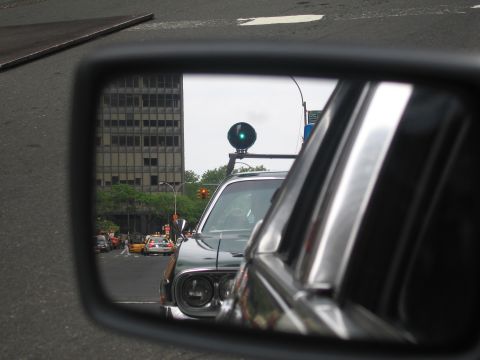 1991 Ford LTD Crown Victoria - car side mirror reflects a green traffic light and a dark vehicle in city traffic, with buildings beyond.