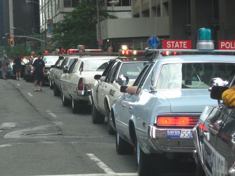 1991 Ford LTD Crown Victoria - long line of light blue and white vintage State Police sedans with roof lights is parked along a city street beneath a red STATE POLICE sig…