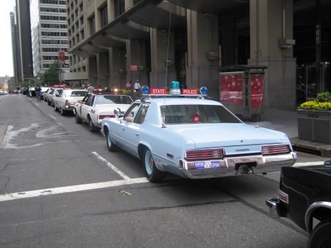 1991 Ford LTD Crown Victoria - light blue vintage two-door coupe is parked at the head of a line of vintage police cars along a downtown curb.