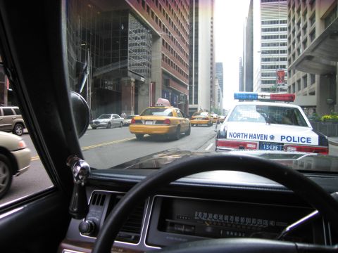 1991 Ford LTD Crown Victoria - Through a windshield over the dash, a North Haven Police sedan and a yellow taxi drive down a downtown avenue lined with towers.
