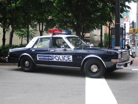 1991 Ford LTD Crown Victoria - Side profile of a dark blue North Haven Police sedan with light bar parked on a city street lined with trees.
