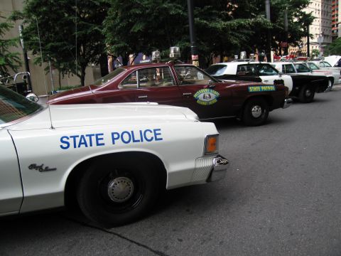 1991 Ford LTD Crown Victoria - lineup of vintage State Police and State Patrol cruisers parked nose-to-tail along a tree-lined downtown street.