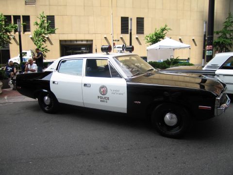 1991 Ford LTD Crown Victoria - Side profile of a black and white vintage LAPD sedan reading 'to protect and to serve' parked on a city street.