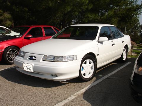 1993 Ford Taurus - A white 1993 Ford Taurus sedan is parked in a lot, with a red car visible to its left.