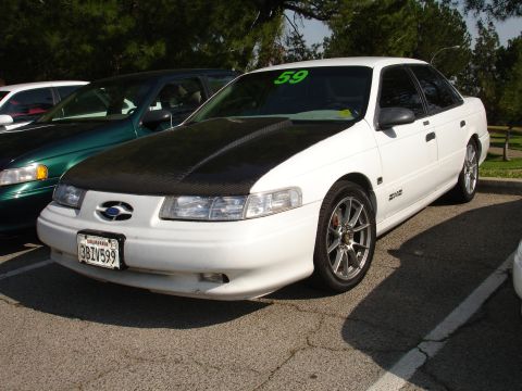 1993 Ford Taurus - A white 1993 Ford Taurus is parked in a lot, showing its aftermarket hood and California license plate.