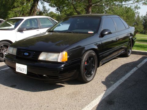 1993 Ford Taurus - A black sedan is parked next to a white car in a lot on a sunny day.