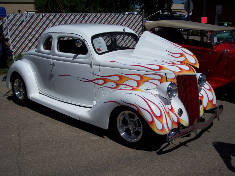 White hot rod coupe with flame graphics and a red grille. Another red car is parked behind it