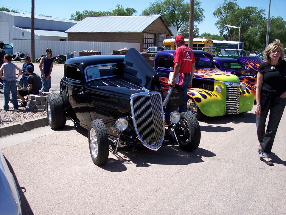 Black hot rod coupe with open hood, parked next to a green and purple truck with flame graphics. People stand around