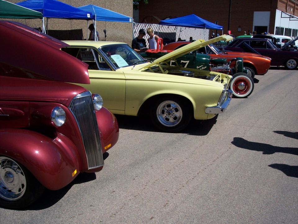 Several cars lined up at a car show, with hoods open. A red car is in the foreground, followed by a yellow car, and then other vehicles