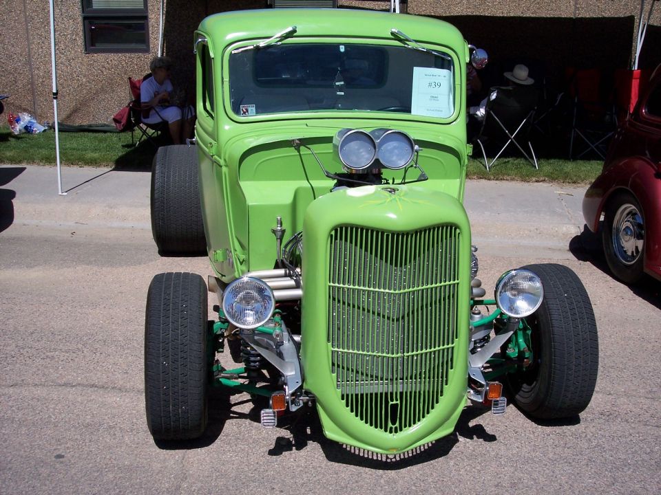 Lime green hot rod truck with prominent vertical grille, dual carburetors, and exposed front suspension. A red car is partially visible to the right
