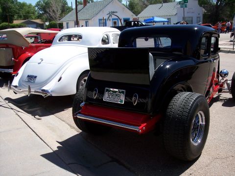 1972 Ford Ranchero - A line of classic cars, including a black hot rod and a white coupe, are parked on a street under a sunny sky.
