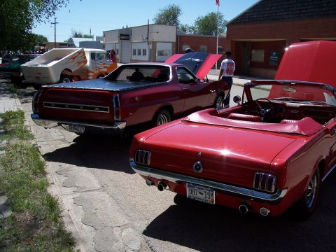 1972 Ford Ranchero - Two classic red cars, a Mustang and a Ranchero, are parked at an outdoor car show with a van in the background.