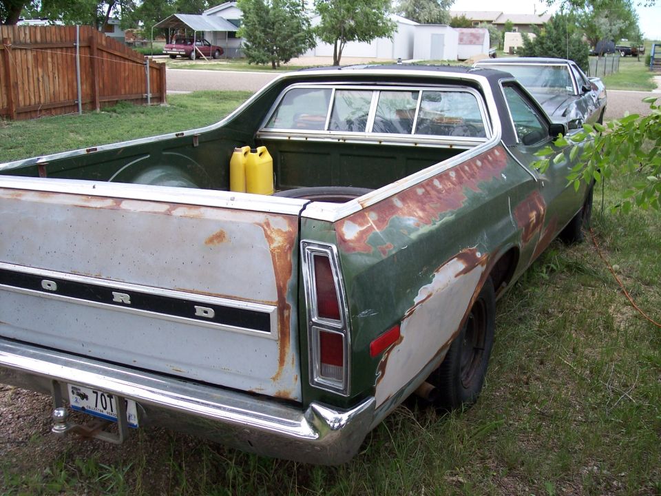 Rear view of a green 1972 Ford Ranchero with rust and primer patches. Two yellow gas cans are in the bed. Another car is parked behind it