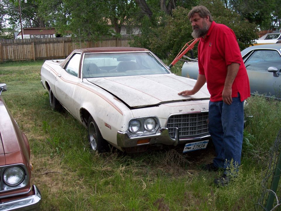 White 1972 Ford Ranchero with orange stripes, hood slightly ajar, parked in grass next to a man