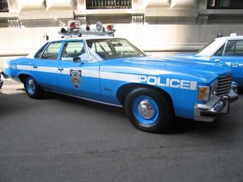 1991 Ford LTD Crown Victoria - Side view of a blue and white vintage NYPD sedan with dual spotlights and roof lights parked on a city street.