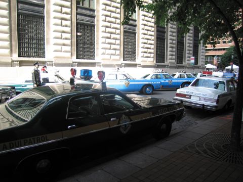1991 Ford LTD Crown Victoria - row of classic patrol cars including a black DUI Patrol sedan and blue-and-white police cruisers parked along a stone building.