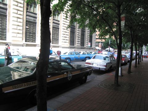 1991 Ford LTD Crown Victoria - tree-lined city street with multiple vintage police cars parked along the curb beside a large stone building.