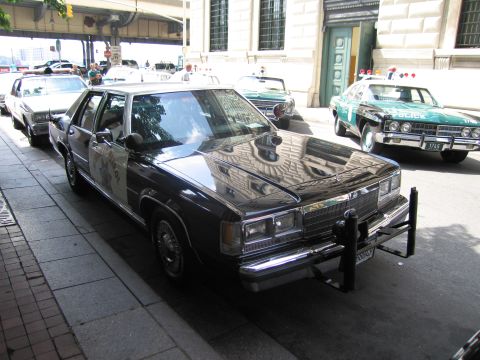 1991 Ford LTD Crown Victoria - black-and-white 1980s Ford police sedan parked at the curb with other classic patrol cars lined up behind it.