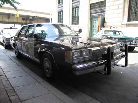 1991 Ford LTD Crown Victoria - dark full-size 1980s sedan fitted with a front push bar parked at the curb among other vintage police cars.