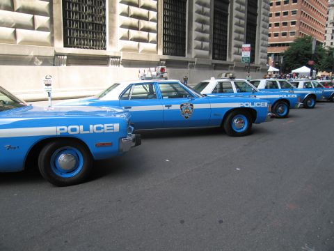 1991 Ford LTD Crown Victoria - line of vintage blue-and-white POLICE sedans with shield emblems parked along a stone building.