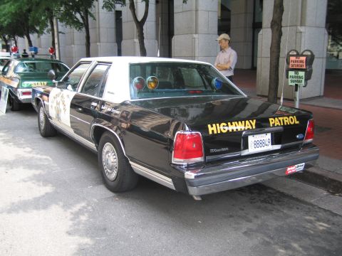 1991 Ford LTD Crown Victoria - Rear view of a black-and-white Ford Highway Patrol sedan with a gold star emblem and a man standing nearby on the sidewalk.
