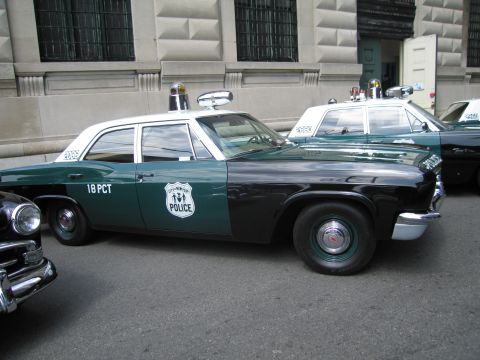 1991 Ford LTD Crown Victoria - Side profile of a green-and-white 18 PCT police sedan with roof beacons and shield decal parked among other classic cruisers.