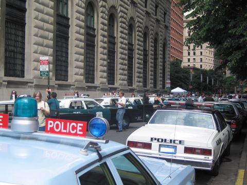 1991 Ford LTD Crown Victoria - busy street scene with several vintage POLICE cars and bystanders gathered beside a large stone building.
