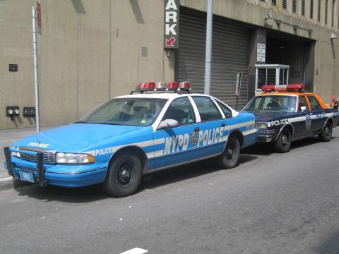 1991 Ford LTD Crown Victoria - blue-and-white NYPD POLICE sedan with a roof light bar parked at the curb ahead of an orange-and-blue cruiser.