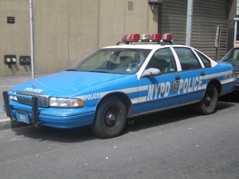 1991 Ford LTD Crown Victoria - blue-and-white NYPD POLICE sedan with a red roof light bar and front push bumper parked along a concrete wall.