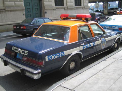 1991 Ford LTD Crown Victoria - Rear quarter view of a navy-and-orange POLICE sedan numbered 9798 PSA 2 with a roof light bar on a city street.