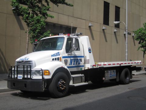 1991 Ford LTD Crown Victoria - white NYPD flatbed tow truck with blue striping, roof beacons and a heavy front push bumper parked beside a building.