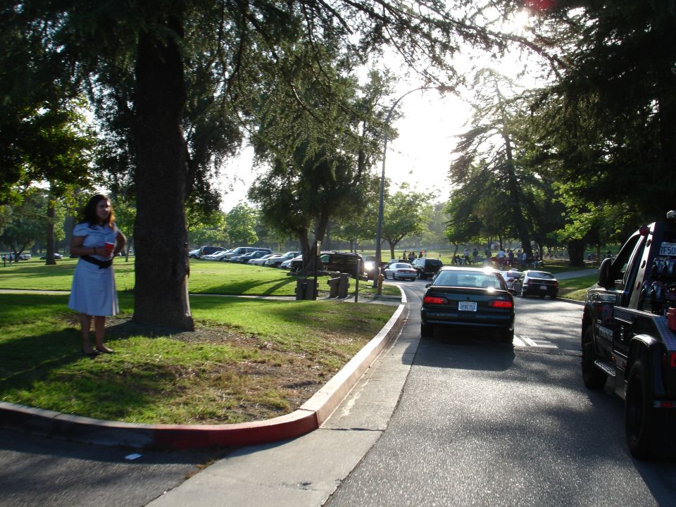 Tow truck on right, cars driving on road, woman standing by tree on left, parked cars in background