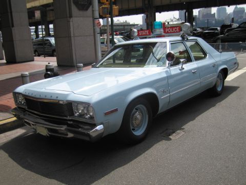 1991 Ford LTD Crown Victoria - light blue vintage State Police four-door sedan with a single rooftop light sits at a waterfront curb beneath an overpass.