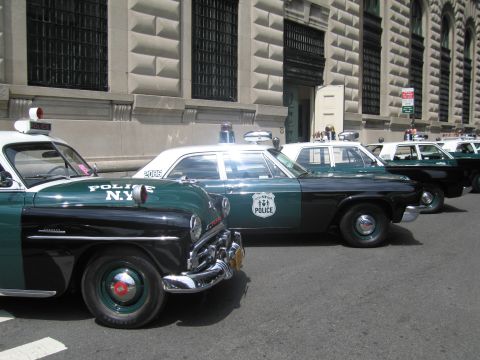 1991 Ford LTD Crown Victoria - lineup of vintage NYPD patrol cars, led by a green-and-black 1950s-style sedan with POLICE N.Y. lettering, along a stone building.
