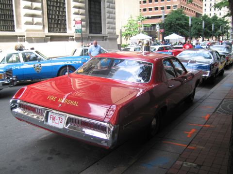 1991 Ford LTD Crown Victoria - red Fire Marshal sedan with plate FM-73 sits parked among vintage police cars on a tree-lined downtown street.