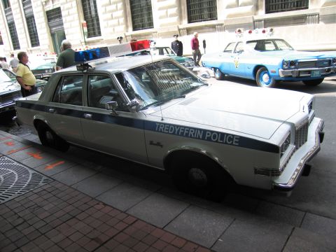 1991 Ford LTD Crown Victoria - white-and-blue Tredyffrin Police sedan with rooftop lightbar is parked curbside among other patrol cars by a stone building.