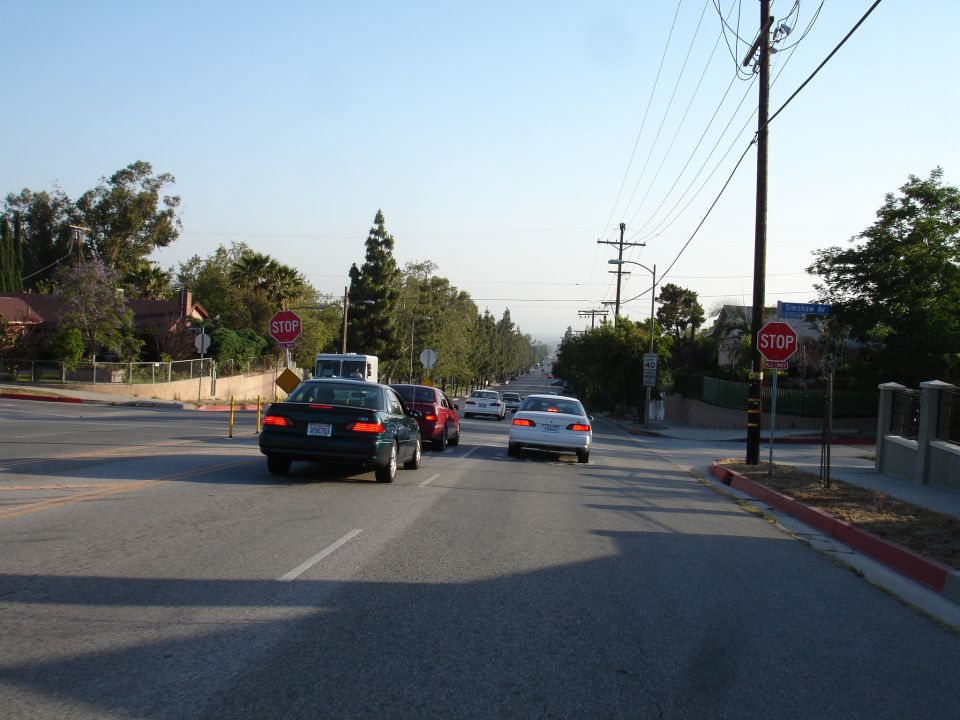 Dark green sedan from rear, driving on a street with other cars and stop signs