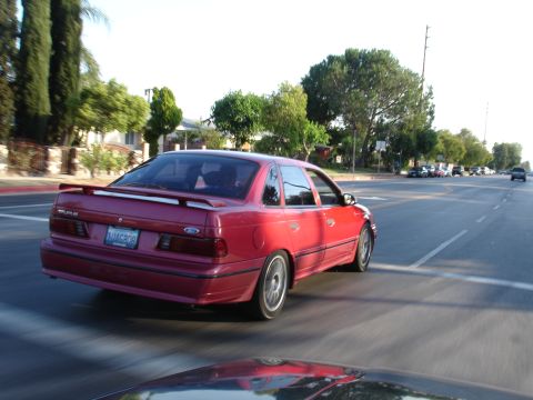 1993 Ford Taurus - A red sedan is captured from behind as it drives down a suburban street.