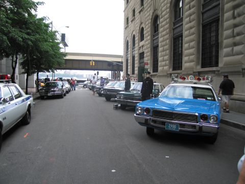 1991 Ford LTD Crown Victoria - downtown street lined with parked vintage police cars, led by a blue sedan bearing the NYC Police plate 2900.