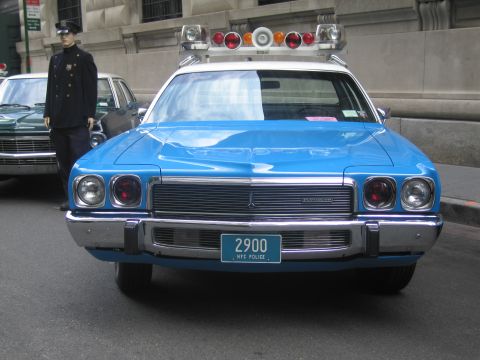 1991 Ford LTD Crown Victoria - Front view of a blue vintage police sedan with rooftop lights and an NYC Police plate 2900, a uniformed officer standing alongside.