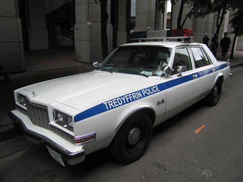 1991 Ford LTD Crown Victoria - white Tredyffrin Police sedan with a blue stripe and single rooftop light sits parked beside a city building.