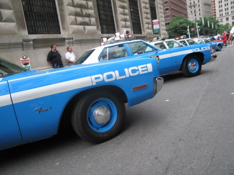 1991 Ford LTD Crown Victoria - tight row of blue-and-white NYPD sedans with POLICE lettering and rooftop lights stretches along a downtown stone building.