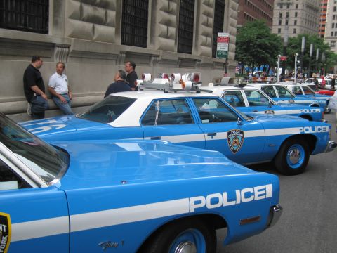 1991 Ford LTD Crown Victoria - Several blue-and-white NYPD patrol cars are lined up curbside as men chat on the sidewalk beside a stone building.