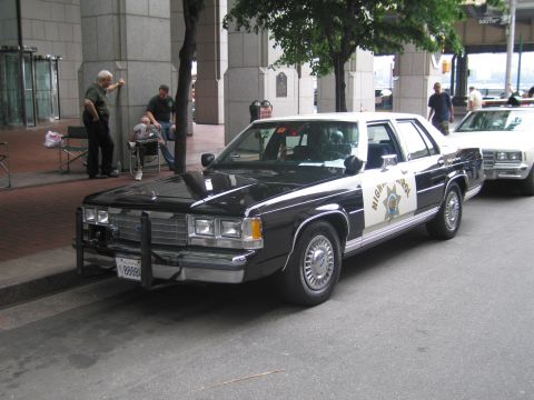 1991 Ford LTD Crown Victoria - black-and-white Highway Patrol sedan with front push bar sits curbside while two men relax in chairs on the sidewalk.