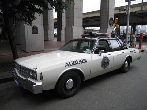 1991 Ford LTD Crown Victoria - white Auburn Police sedan with rooftop lightbar and door shield is parked curbside near an elevated roadway.