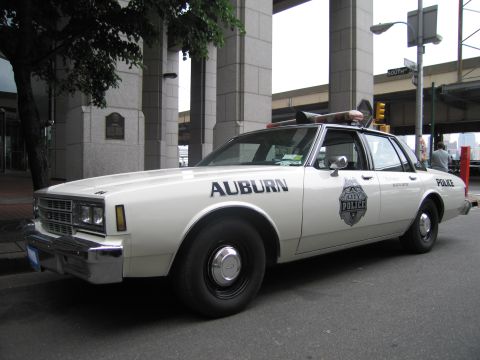 1991 Ford LTD Crown Victoria - white Auburn Police sedan with rooftop lights and a POLICE door shield sits parked along a downtown curb near an overpass.