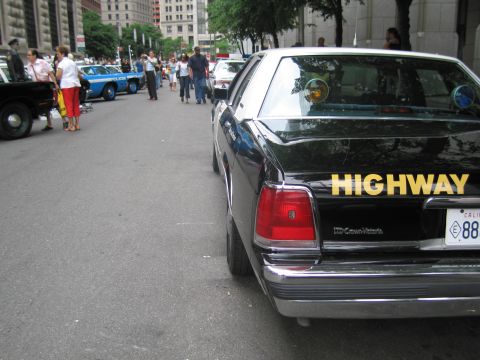 1991 Ford LTD Crown Victoria - Rear of a black Crown Victoria patrol car with yellow HIGHWAY lettering and a California plate, parked on a busy city street.