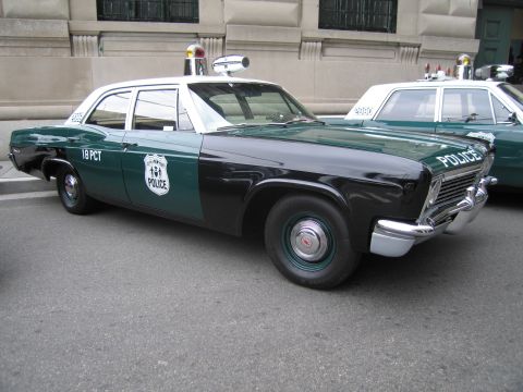 1991 Ford LTD Crown Victoria - green-and-black police sedan lettered '18 PCT' with roof beacons among a row of vintage NYPD-style patrol cars by a building.