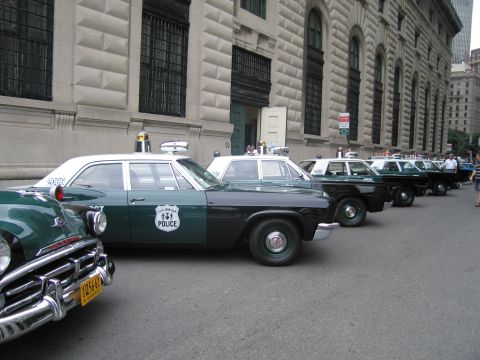 1991 Ford LTD Crown Victoria - long line of vintage green-and-black police cars with roof beacons parked diagonally beside a large stone building, pedestrians nearby.